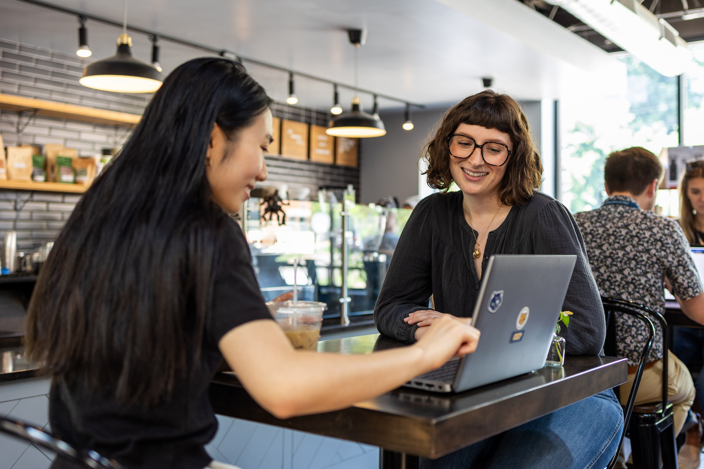Two students sitting at a table in a coffee shop looking at a laptop together.
