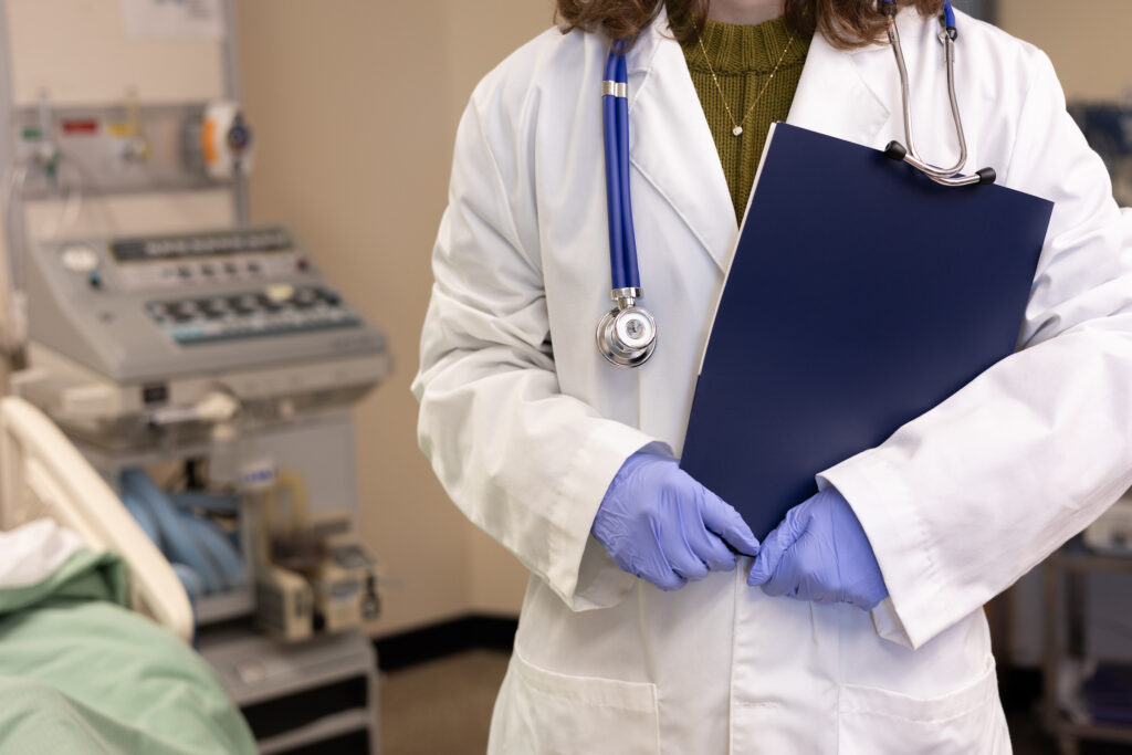 Person wearing a white lab coat wearing a stethoscope and holding a blue notebook.