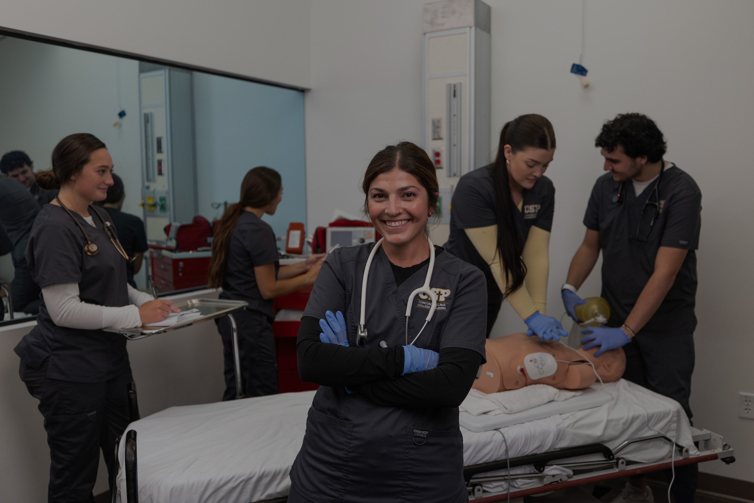 Nursing student wearing a stethoscope and gloves standing in a lab setting in front of other nursing students who are working on a simulation.